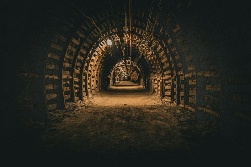 A dimly lit underground mine tunnel receding into darkness, a single lamp glowing in the distance