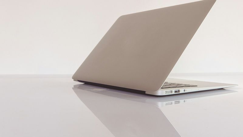 A silver MacBook Air seen from behind, its lid open, on a white table that reflects it