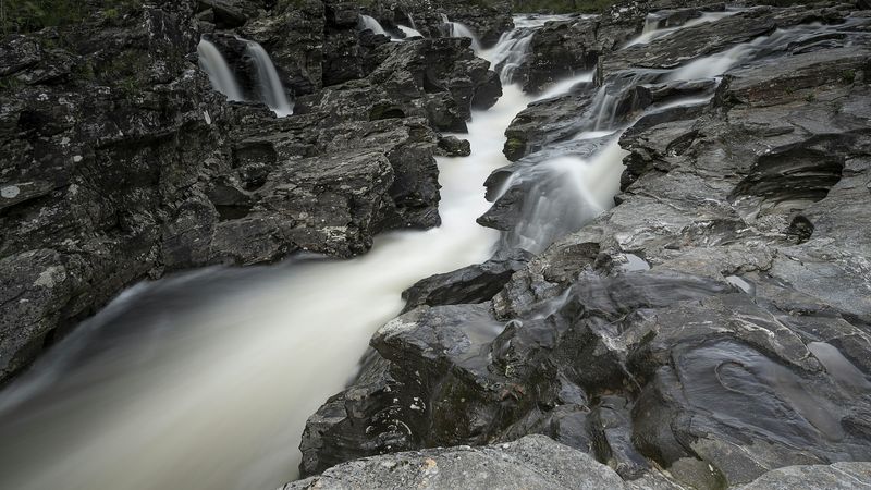 A long-exposure photograph of a river forcing its way through a channel of dark rock — the water never stops moving, finding a path around every obstruction