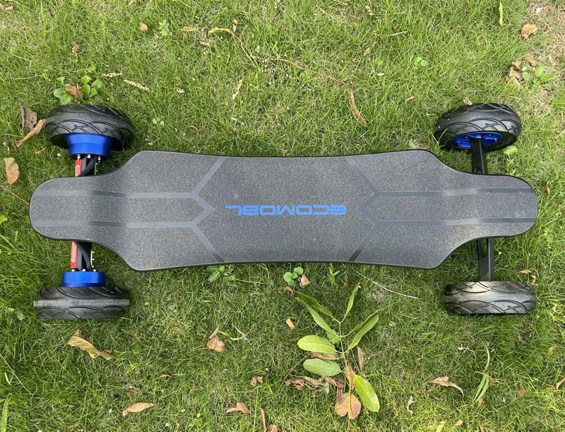 An all-terrain electric longboard photographed from directly above, lying on grass — griptape deck, pneumatic tyres and blue motor hubs.
